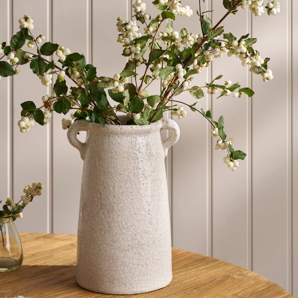 White textured vase with greenery on a wooden surface against a light gray wall.
