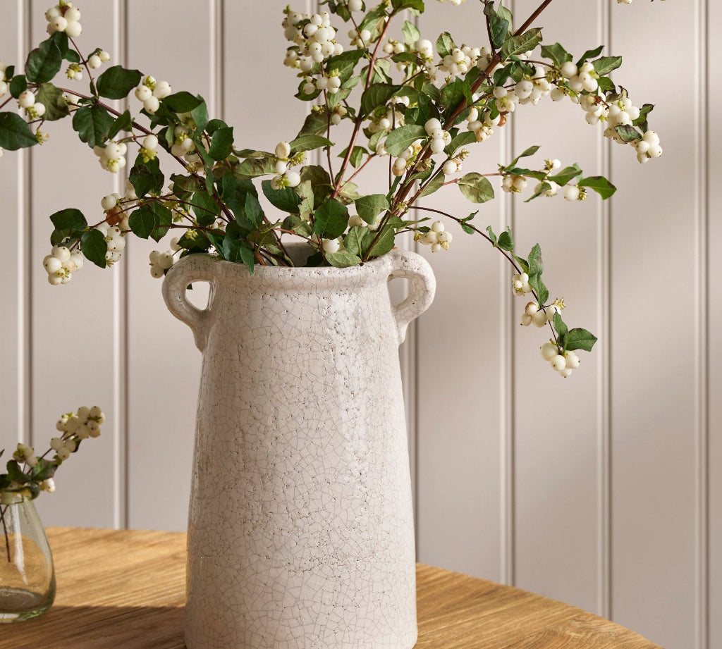 White textured vase with greenery on a wooden surface against a light gray wall.