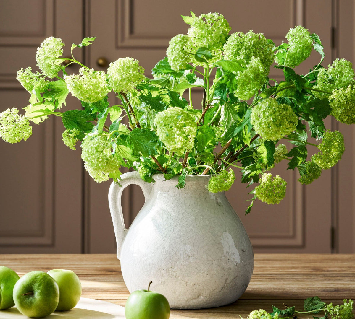 White pitcher with green flowers on a wooden table with apples
