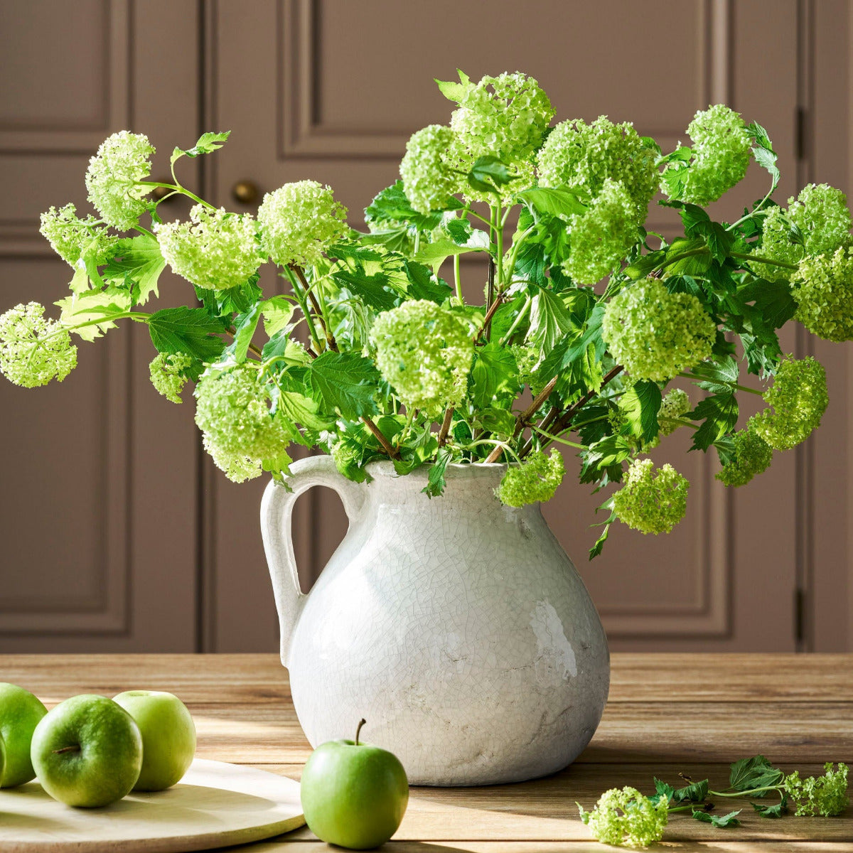 White pitcher with green flowers on a wooden table with apples