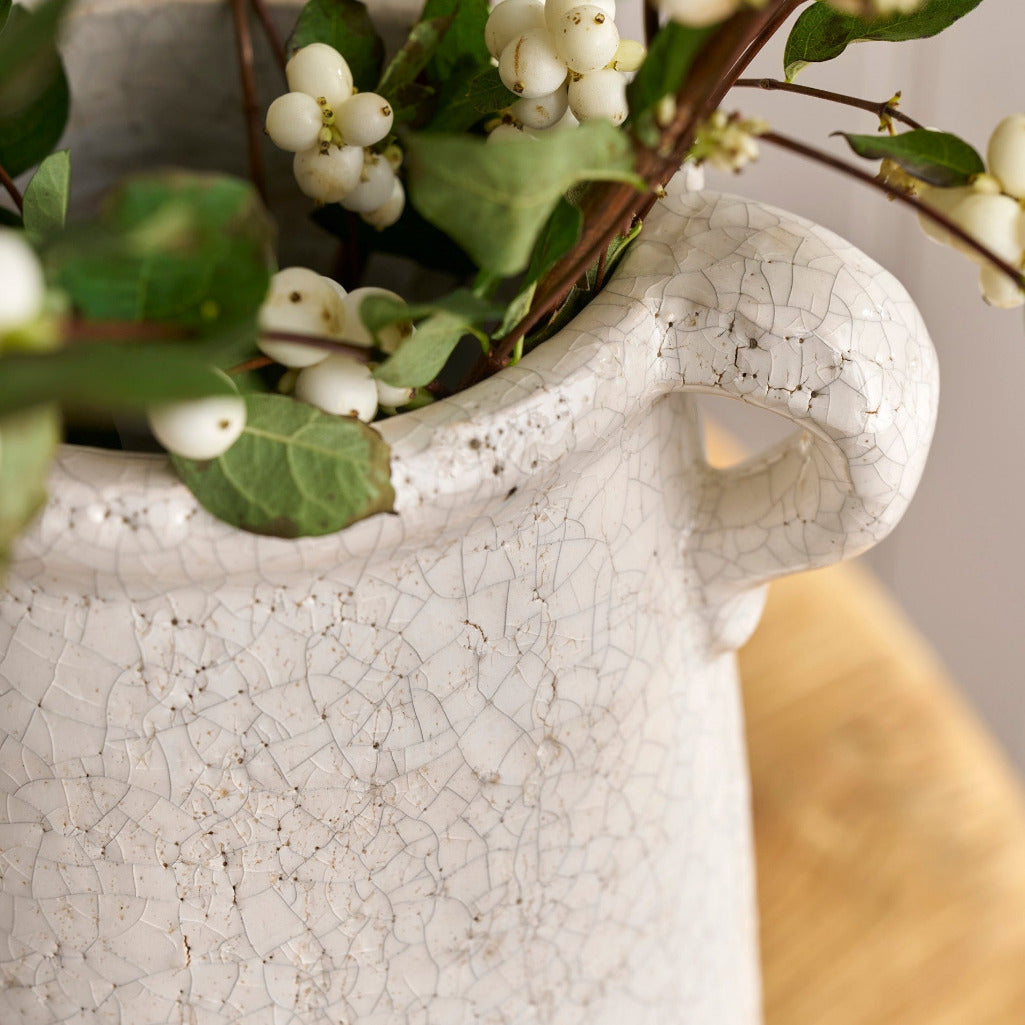 White ceramic vase with cracked texture holding greenery and white flowers on a wooden surface.
