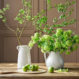 Two white ceramic vases with green branches and apples on a wooden surface against a brown paneled wall.