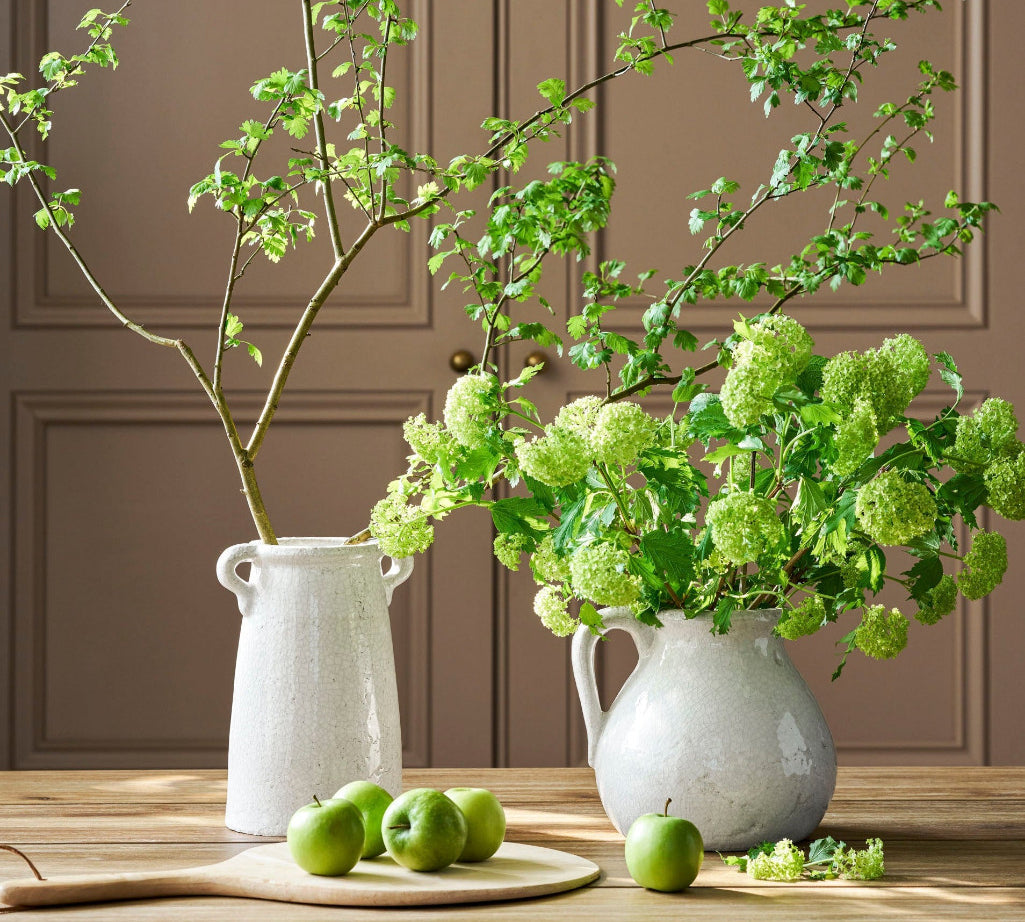 Two white ceramic vases with green branches and apples on a wooden surface against a brown paneled wall.