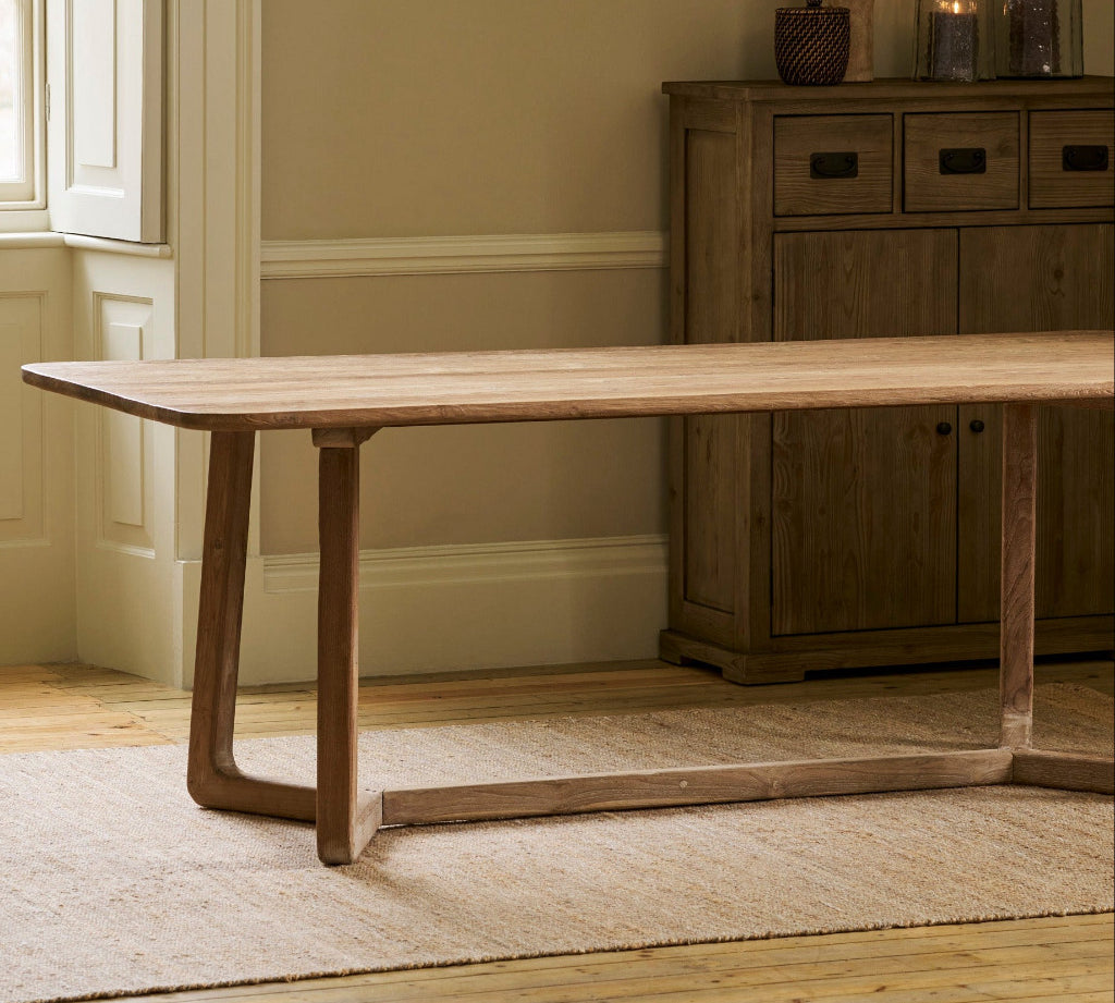 A light natural reclaimed teak wood dining table with a unique angled top and curved legs, styled in a warm, neutral-toned room next to a wooden sideboard.