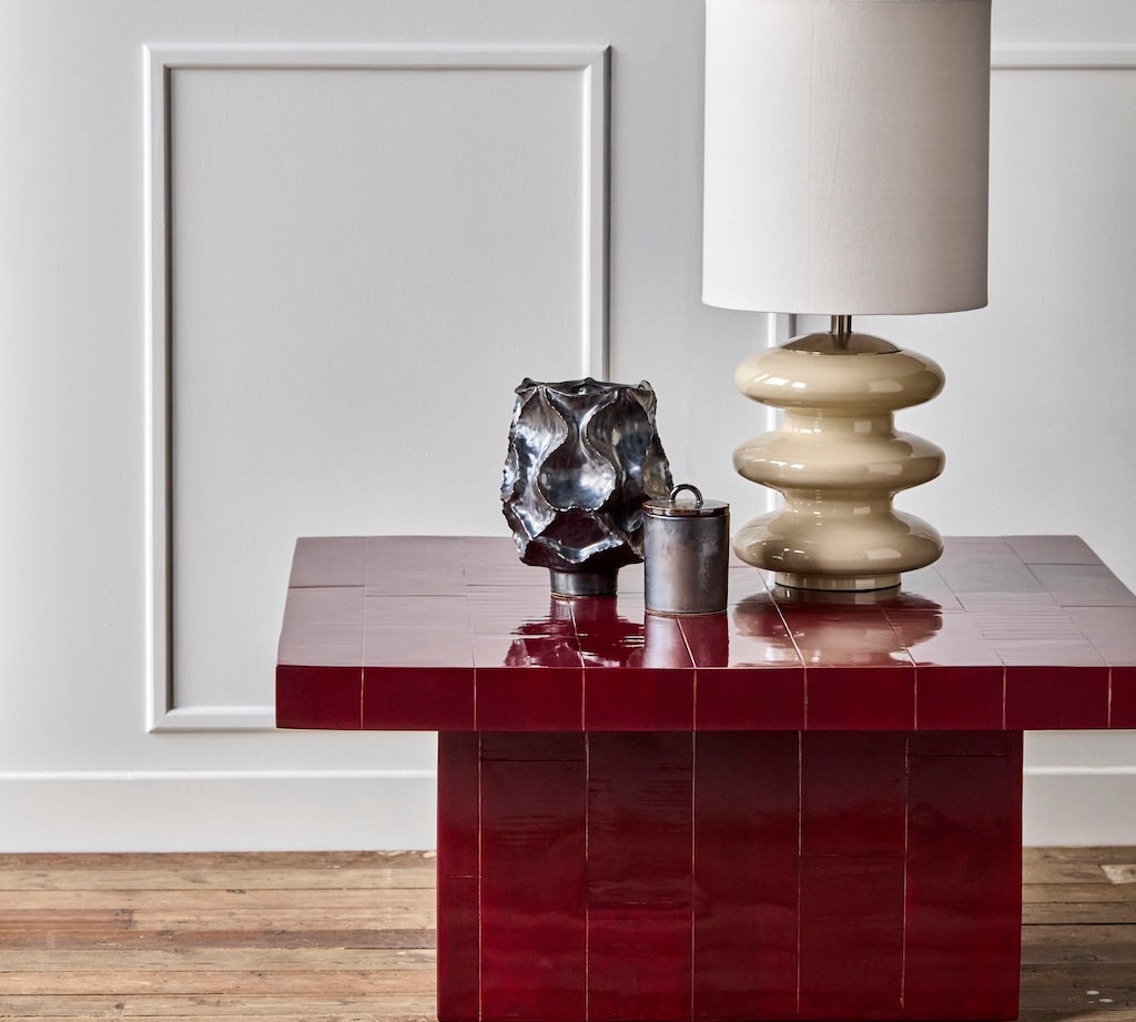 A glossy deep red tiled side table with a tiered cream ceramic table lamp and two decorative vases on top, set against a white paneled wall and light wood floor.