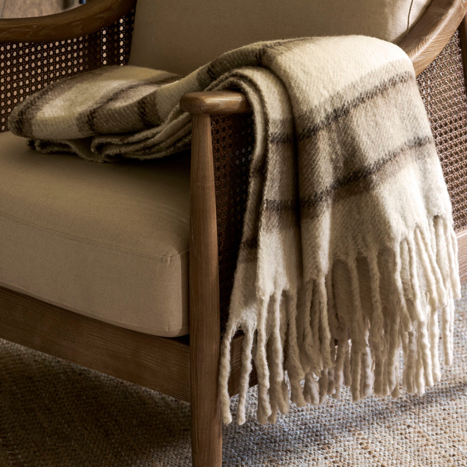 A mid-century modern armchair with beige cushions, a woven cane backrest, and a cream throw blanket draped over the arm, set on a textured rug in a warm interior.