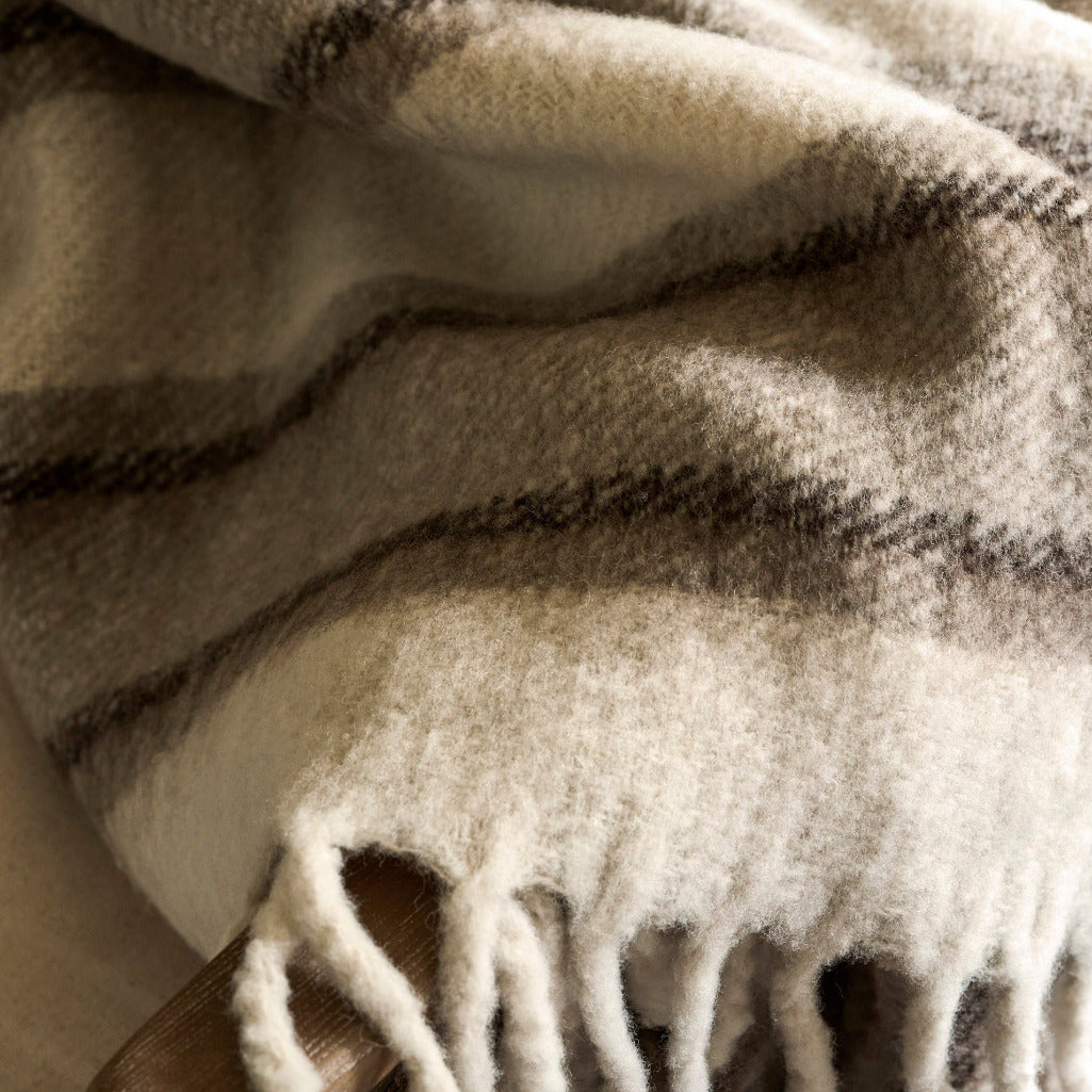 A close-up of a soft woven throw blanket with fringe, draped in folds. The blanket has a warm cream base with dark brown and gray plaid stripes.