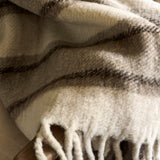 A close-up of a soft woven throw blanket with fringe, draped in folds. The blanket has a warm cream base with dark brown and gray plaid stripes.