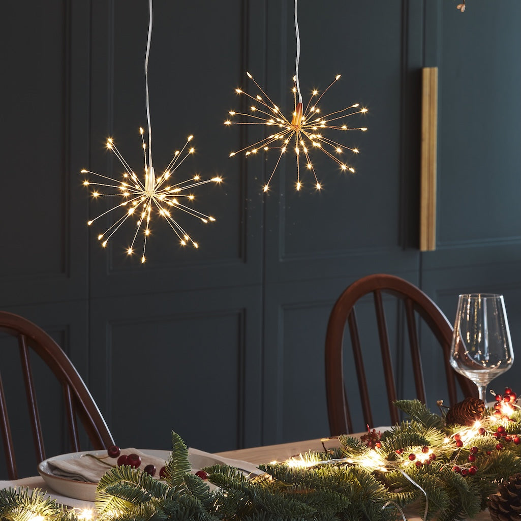 Two spherical starburst LED pendant lights with warm white lights hanging above a decorated dining table with pine garlands and a wine glass, set against a dark paneled wall.