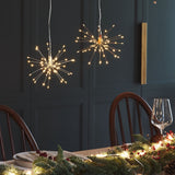 Two spherical starburst LED pendant lights with warm white lights hanging above a decorated dining table with pine garlands and a wine glass, set against a dark paneled wall.
