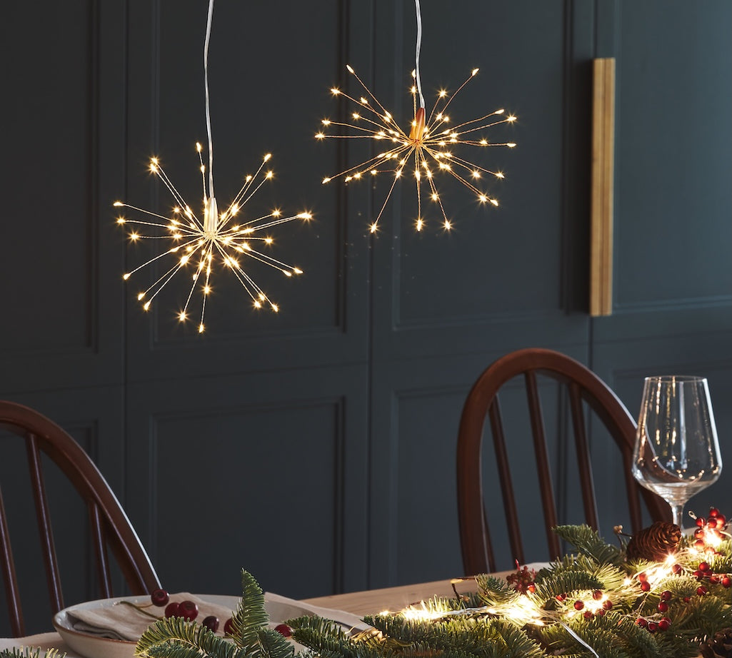 Two spherical starburst LED pendant lights with warm white lights hanging above a decorated dining table with pine garlands and a wine glass, set against a dark paneled wall.