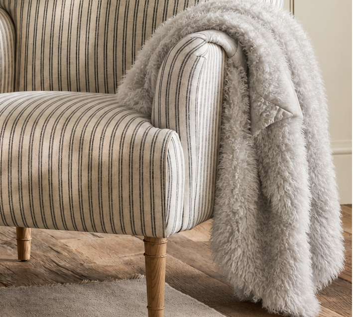 A classic armchair with beige and gray vertical stripes, styled with a fluffy light gray throw blanket draped over one arm, set in a warm, neutral-toned living room.