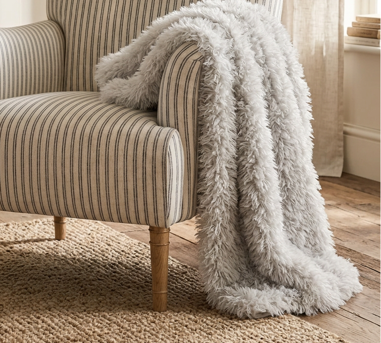 A classic armchair with beige and gray vertical stripes, styled with a fluffy light gray throw blanket draped over one arm, set in a warm, neutral-toned living room.