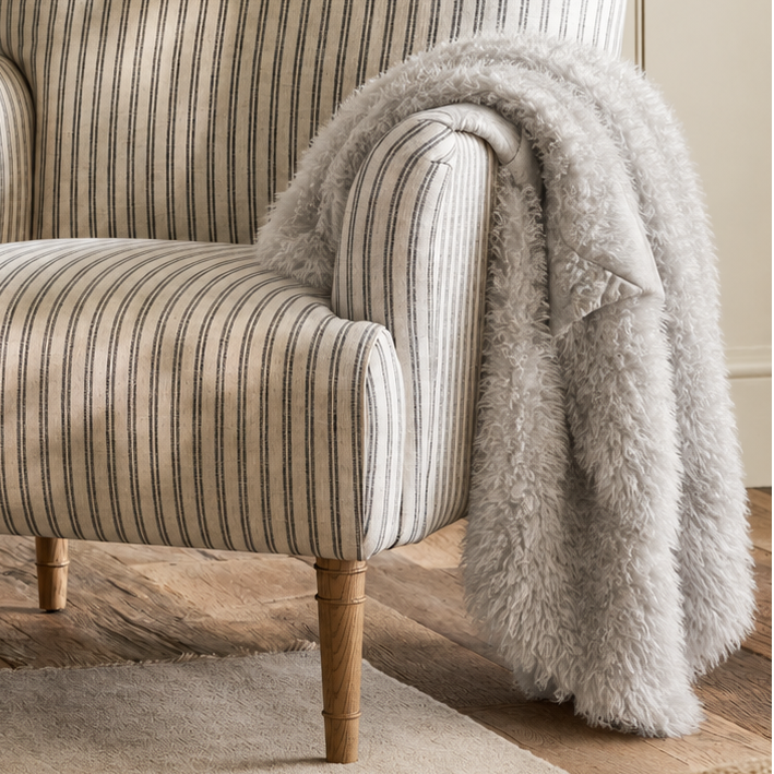 A classic armchair with beige and gray vertical stripes, styled with a fluffy light gray throw blanket draped over one arm, set in a warm, neutral-toned living room.