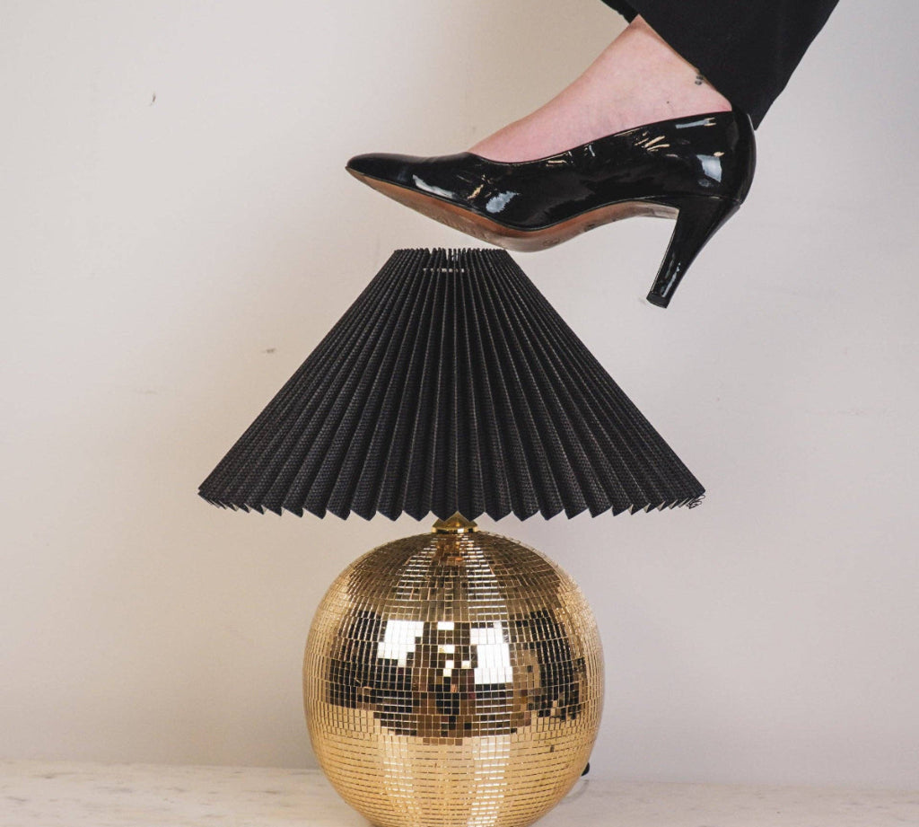 Gold disco ball table lamp with black pleated shade and high-heeled foot stepping on it.
