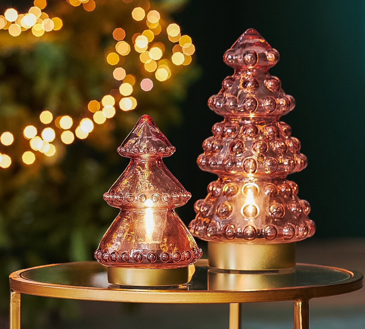 Two pink glass Christmas tree lights on a gold table with blurred lights in the background
