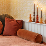 A cozy bedroom corner with patterned wallpaper, a bed dressed in rust and peach bedding, and several velvet pillows including a large rust square pillow and a brown cylindrical bolster pillow. A white headboard or shelf holds three lit taper candles in amber and dark glass holders.