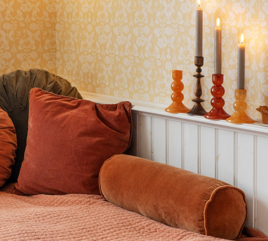 A cozy bedroom corner with patterned wallpaper, a bed dressed in rust and peach bedding, and several velvet pillows including a large rust square pillow and a brown cylindrical bolster pillow. A white headboard or shelf holds three lit taper candles in amber and dark glass holders.