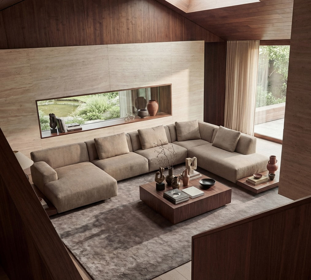 A modern sectional sofa in a warm beige fabric, arranged in an L-shape within a contemporary minimalist living room featuring wood paneling, a large textured gray rug, and a low wooden coffee table.