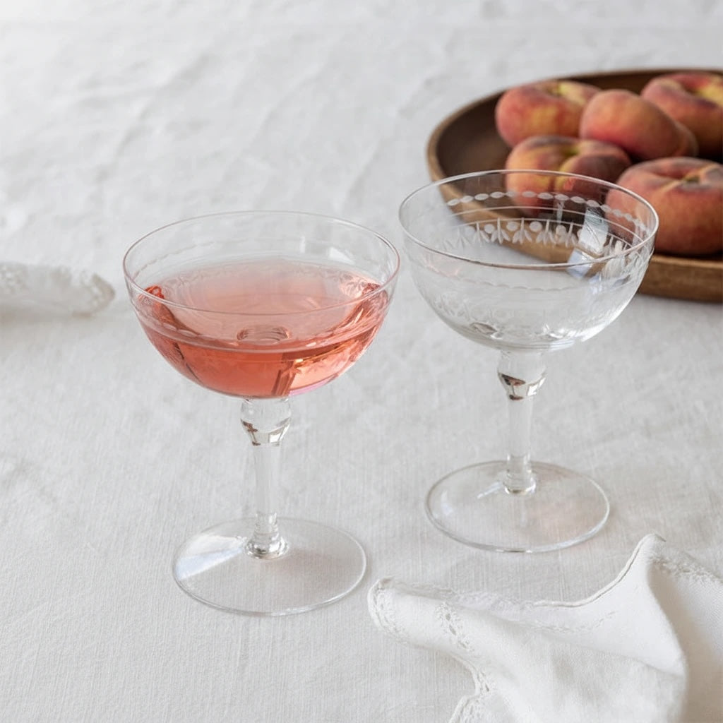 Two clear coupe-style wine glasses on a white fabric surface, one filled with pink liquid and one empty, with peaches blurred in the background.