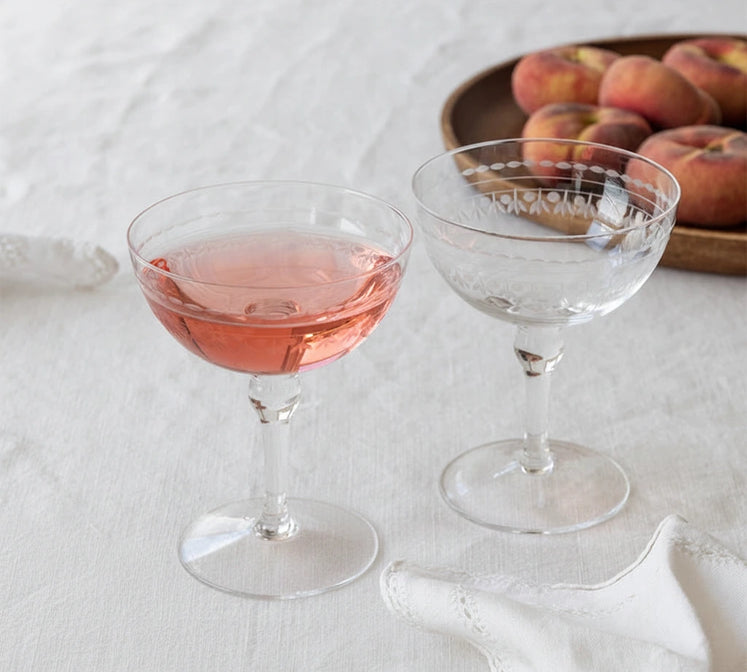 Two clear stemmed coupe glasses on a white tablecloth, one filled with pink liquid and one empty, with a plate of peaches in the background.