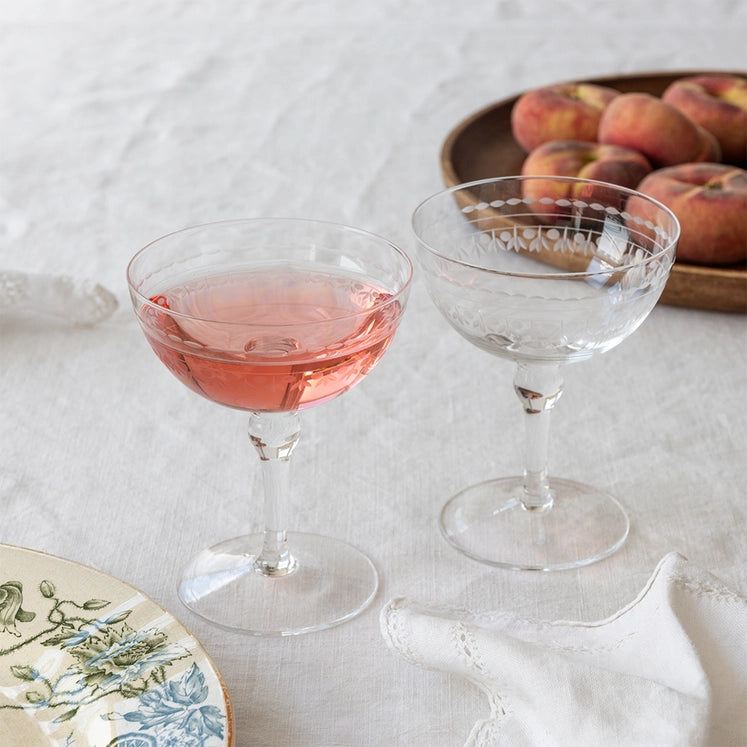 Two clear stemmed coupe glasses on a white tablecloth, one filled with pink liquid and one empty, with a plate of peaches in the background.