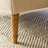 Close-up of a beige upholstered furniture leg with a natural wood tapered post, standing on a textured cream woven rug.