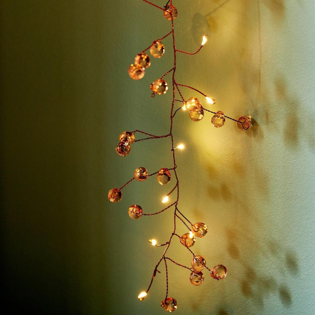 A decorative branch-shaped string light with small warm white bulbs and clear faceted crystal-like beads on a thin red wire, shown hanging against a softly lit wall.