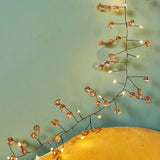 Wire string light garland with clear faceted crystal beads and warm white LED lights, draped across a yellow surface against a teal background.