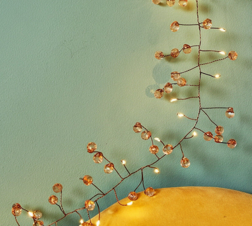 Wire string light garland with clear faceted crystal beads and warm white LED lights, draped across a yellow surface against a teal background.