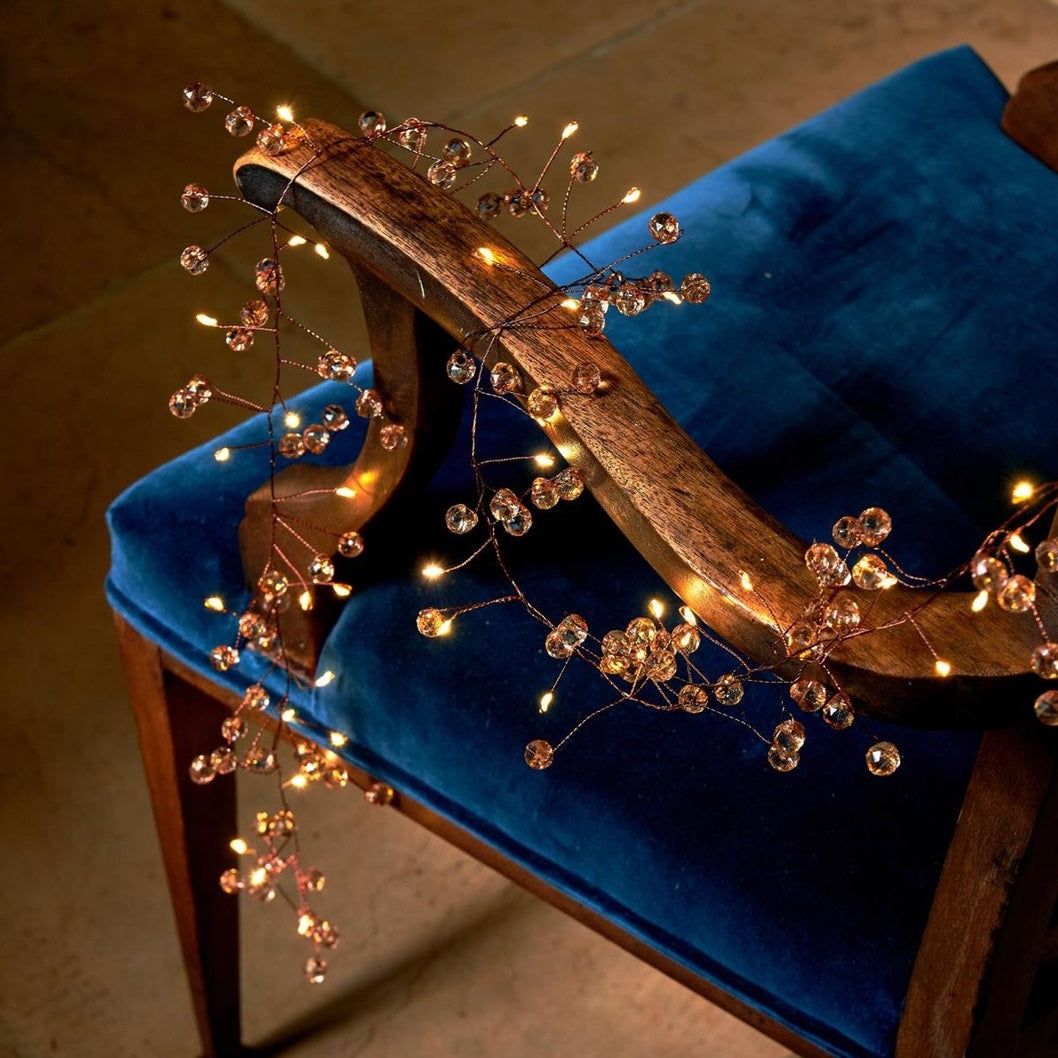 Warm white vine string lights with clear crystal beads draped over a blue velvet upholstered wooden chair in a dimly lit indoor setting.