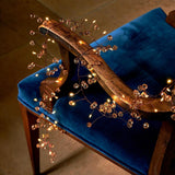 Warm white vine string lights with clear crystal beads draped over a blue velvet upholstered wooden chair in a dimly lit indoor setting.