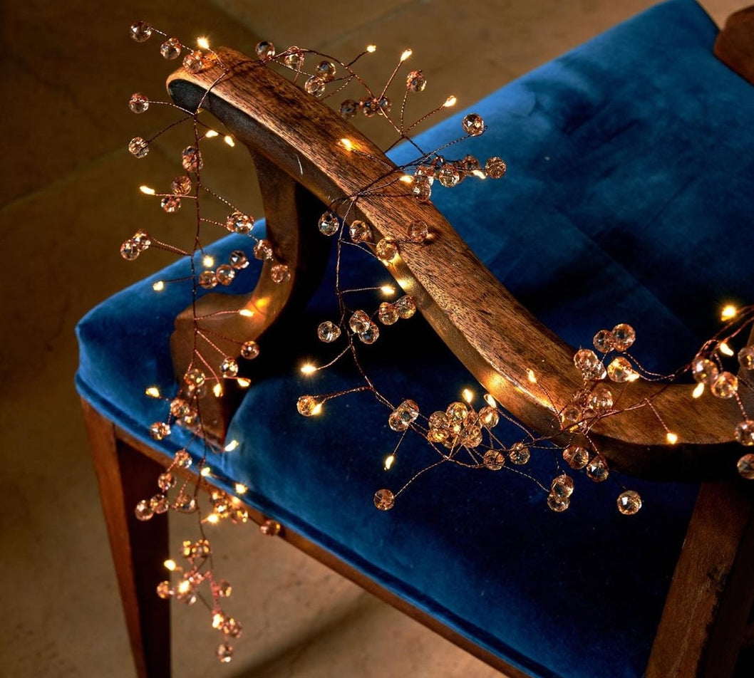 Warm white vine string lights with clear crystal beads draped over a blue velvet upholstered wooden chair in a dimly lit indoor setting.