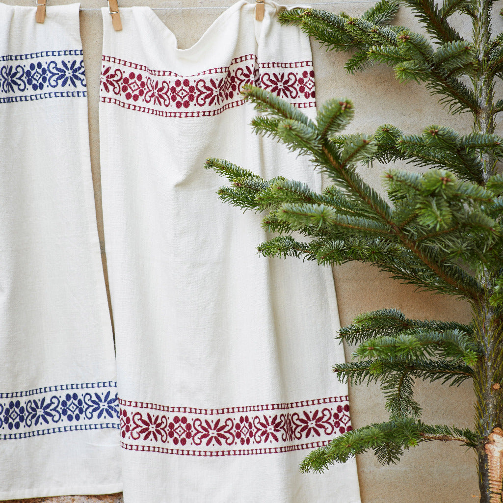 A white linen garment or textile hanging on a wooden clothesline, decorated with embroidered borders in blue and burgundy red floral patterns, styled next to a green pine tree.