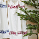 A white linen garment or textile hanging on a wooden clothesline, decorated with embroidered borders in blue and burgundy red floral patterns, styled next to a green pine tree.