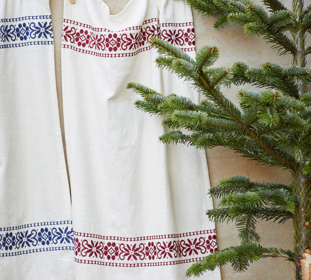 A white linen garment or textile hanging on a wooden clothesline, decorated with embroidered borders in blue and burgundy red floral patterns, styled next to a green pine tree.