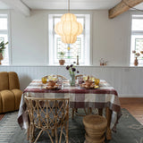 A dining table set in a bright, modern dining room with a red and white checkered tablecloth, woven chairs, a hanging paper lantern pendant light, and a rustic wooden stool on a patterned rug.