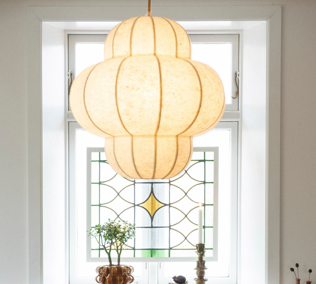 A dining room scene with a table set for two under a large cream paper lantern pendant light. The table has a red-and-cream checkered tablecloth, matching plates, amber glasses, and a vase of dark flowers.