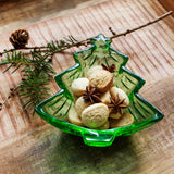 A green Christmas tree-shaped glass bowl filled with cookies and star anise, styled on a rustic wooden tray with pine branches and a pinecone.