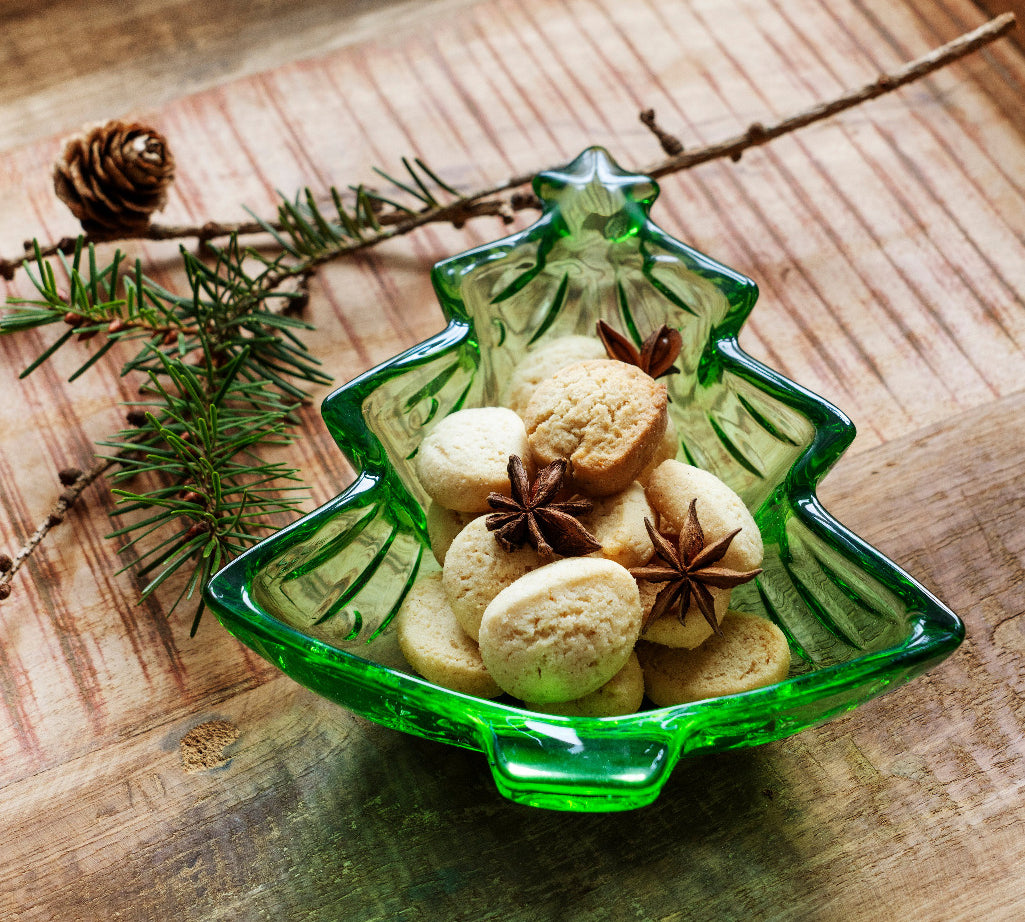 A green Christmas tree-shaped glass bowl filled with cookies and star anise, styled on a rustic wooden tray with pine branches and a pinecone.