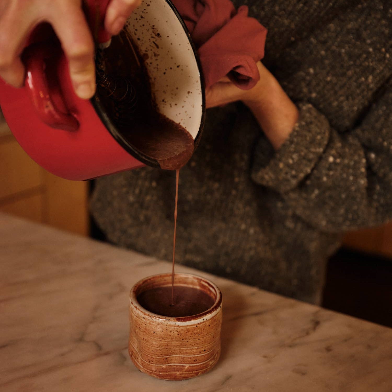 Person pouring hot chocolate from a red container into a small brown cup on a marble surface.