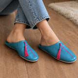 Blue slippers with pink tassels worn by a person on a wooden floor.