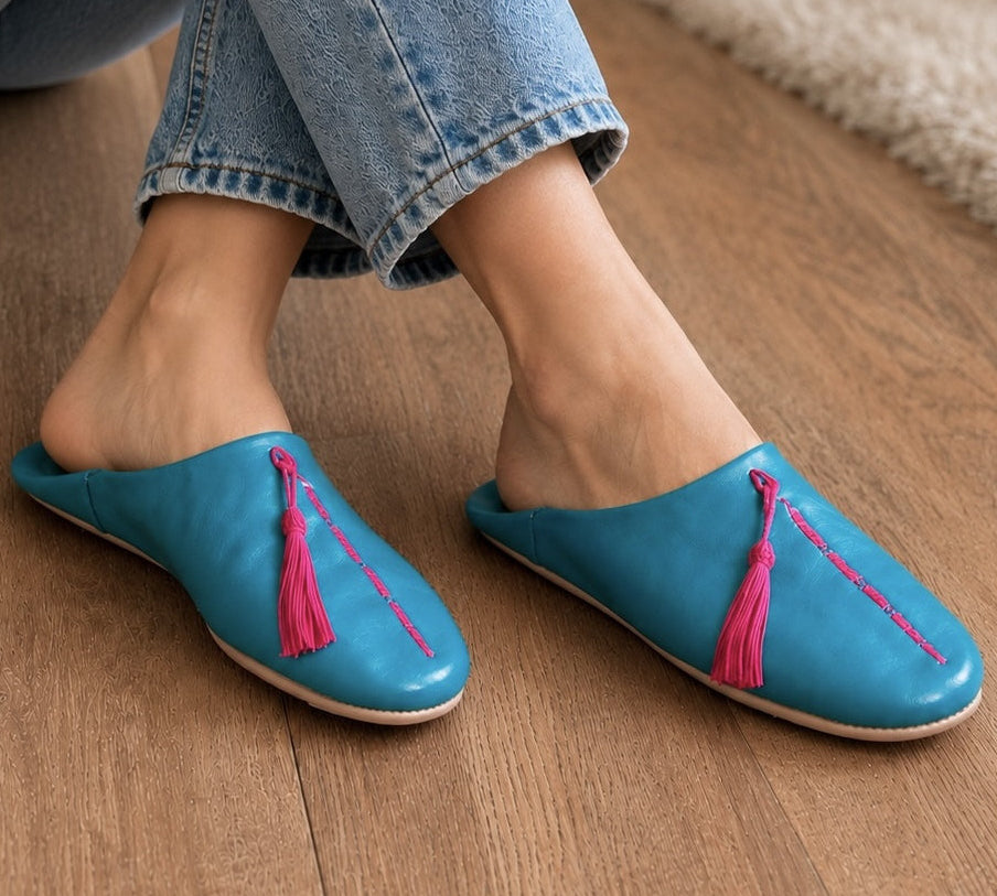 Blue slippers with pink tassels worn by a person on a wooden floor.