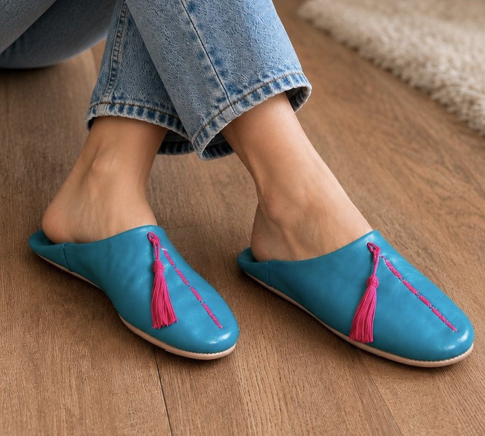 Blue slippers with pink tassels worn by a person on a wooden floor.