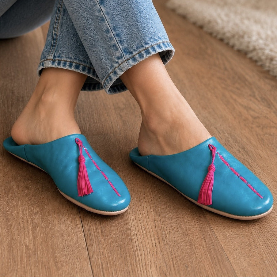 Blue slippers with pink tassels worn by a person on a wooden floor.