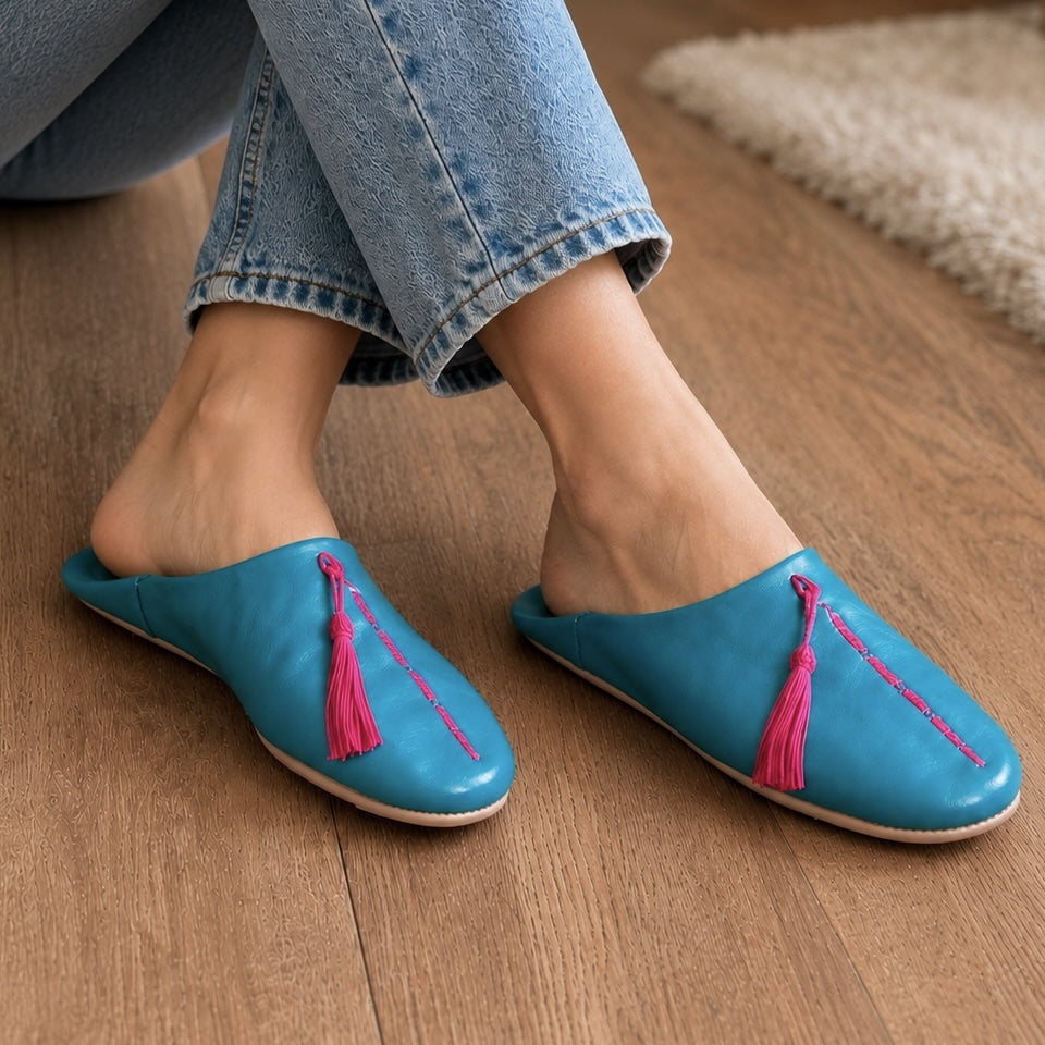 Blue slippers with pink tassels worn by a person on a wooden floor.