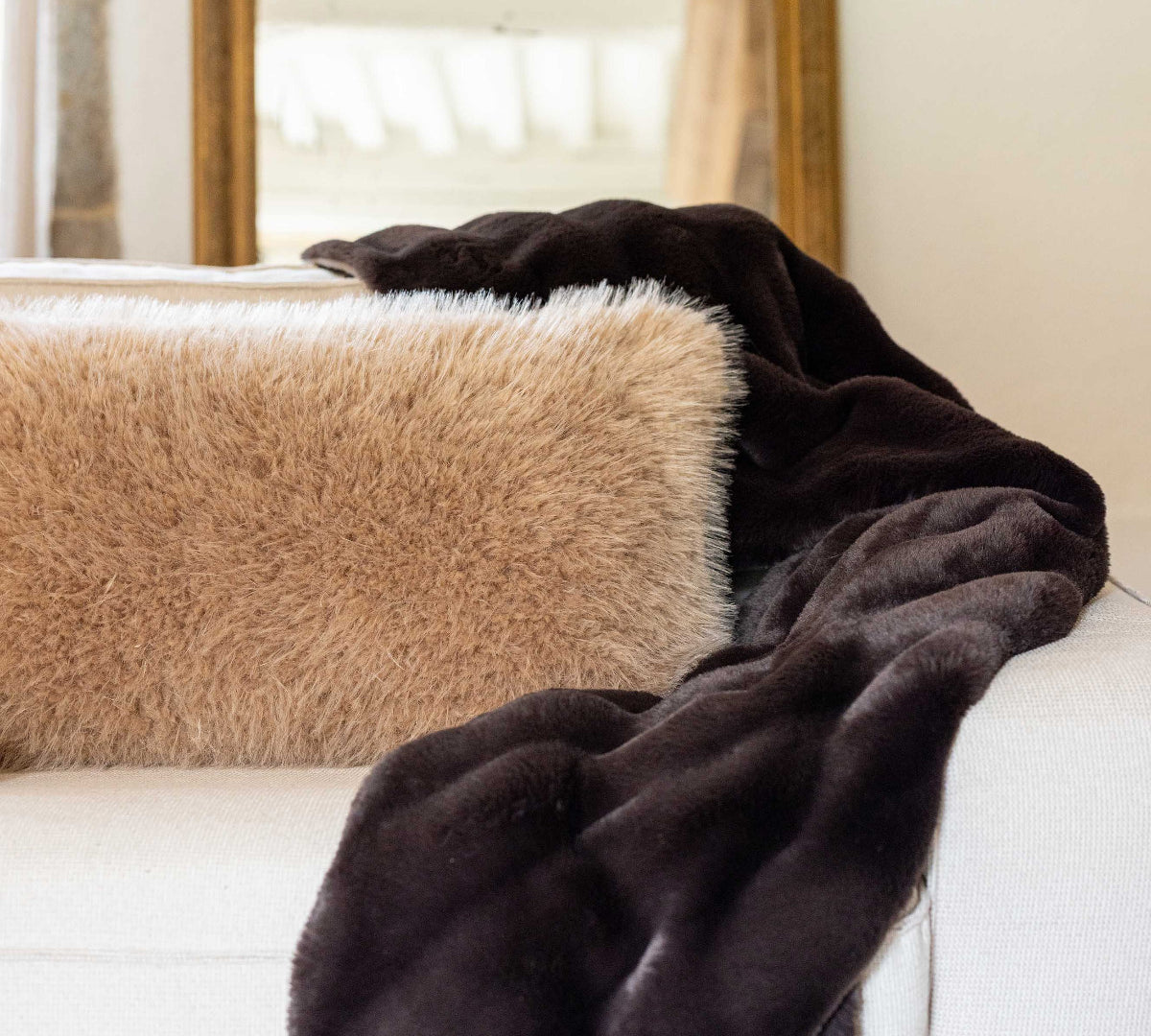 A dark brown plush faux fur throw blanket draped over a white sofa next to a beige shag pillow, with a gold-framed mirror in the background.