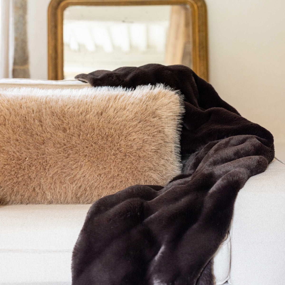 A dark brown plush faux fur throw blanket draped over a white sofa next to a beige shag pillow, with a gold-framed mirror in the background.