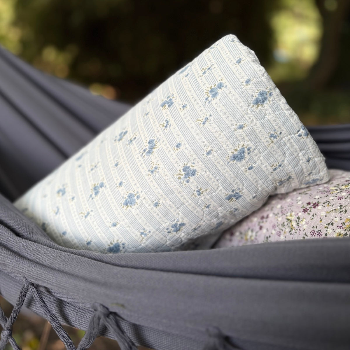 A light blue and white striped quilted pillow with small blue floral motifs rests on a gray hammock outdoors, with another floral pillow partially visible underneath.