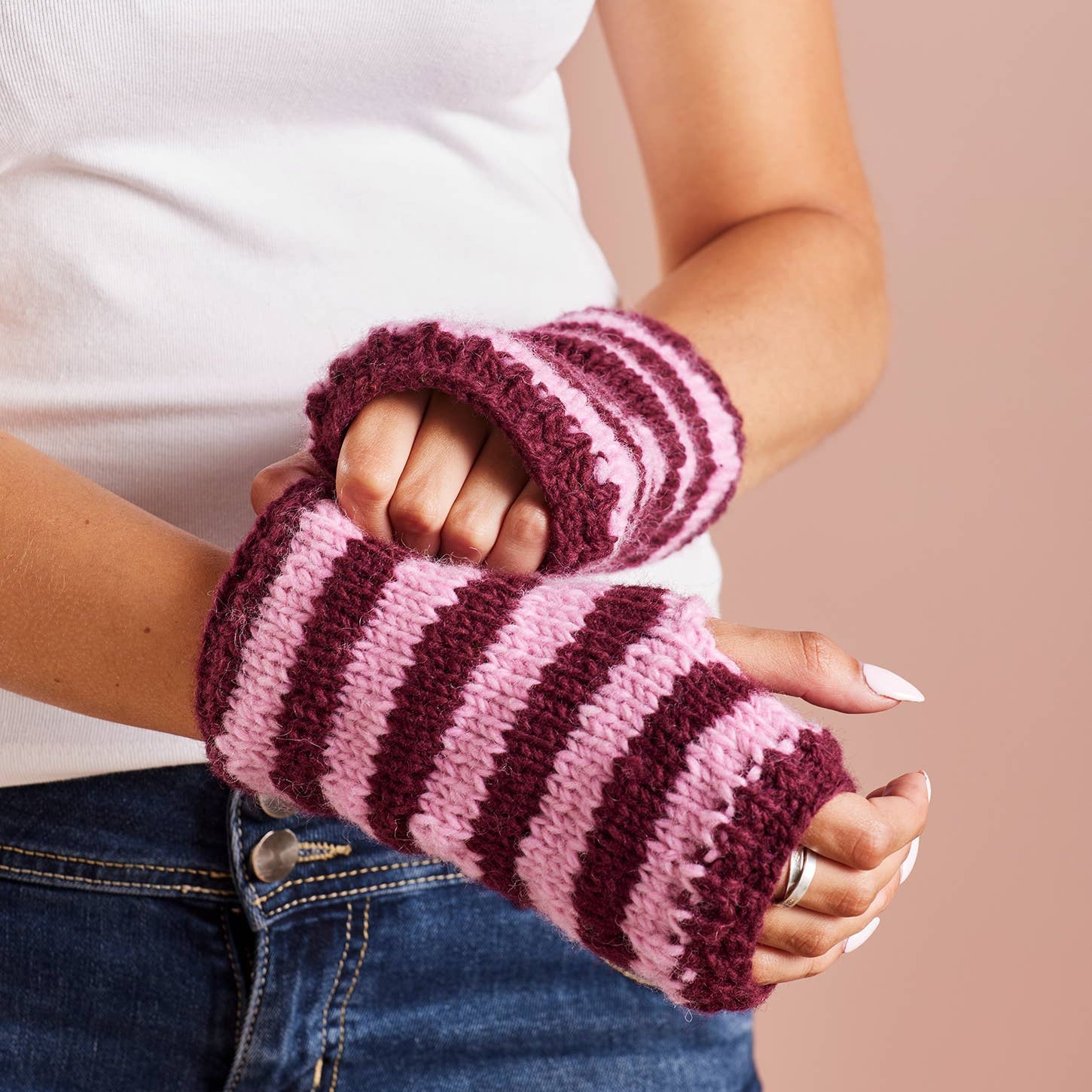 A person wearing pink and burgundy striped knit fingerless wrist warmers, paired with a white t-shirt and blue jeans, against a solid dusty pink background.